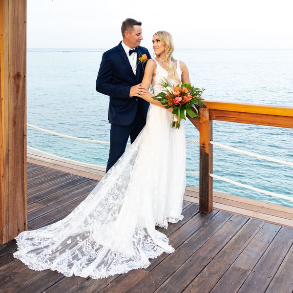 Bride in White Wedding Dress With Hair Down Holding Tropical Bouquet Posing with Groom in Navy Tuxedo by The Ocean