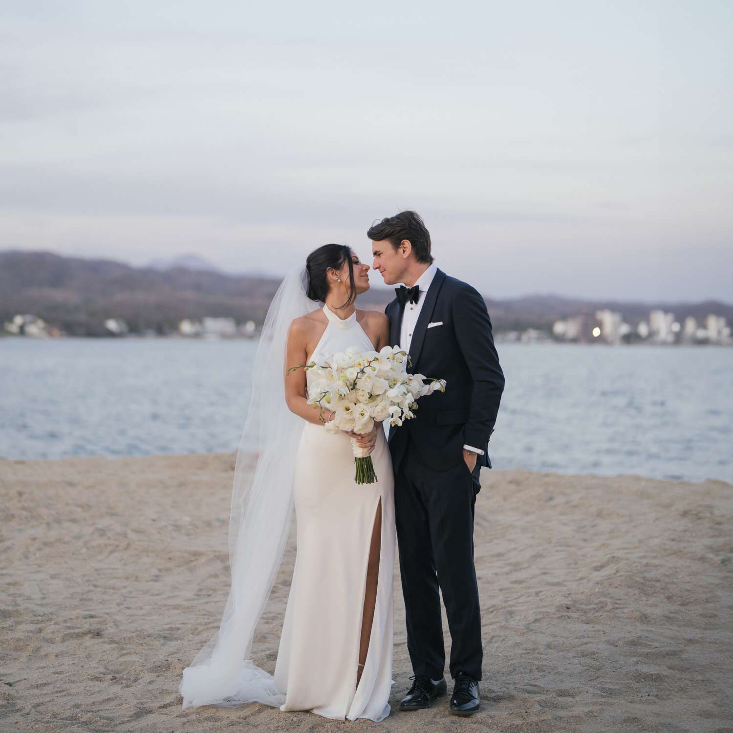 bride and groom couple portrait on sandy beach holding flower bouquet leaning in for a kiss