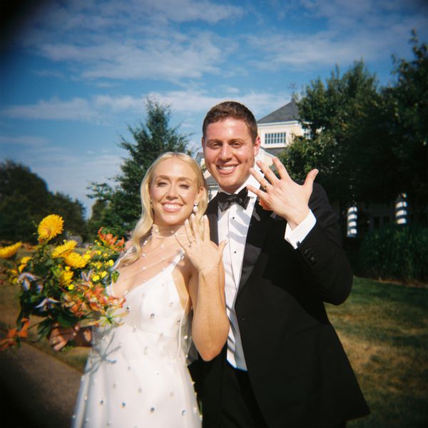 Bride in White Wedding Dress With Pearl Studs Posing With Groom in Black Tuxedos Holding Up Hands With Wedding Rings