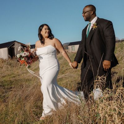 Bride in Strapless Wedding Dress Holding Hands With Groom in Black Suit With Teal Tie in Grassy Field