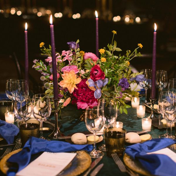 Round tables with dark green velvet linens, purple taper candles, gold chargers, and royal blue napkins