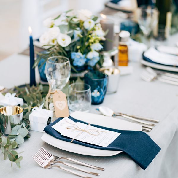 A navy blue napkin and a white menu tied together with a string on top of a white plate set on a table with white linens, greenery, white flowers, and blue taper candles