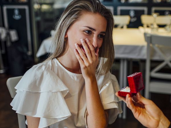 woman in a white shirt gets emotional and covers her mouth with her hand while her boyfriend takes her hand and proposes to her in a restaurant while offering her a ring in a red box