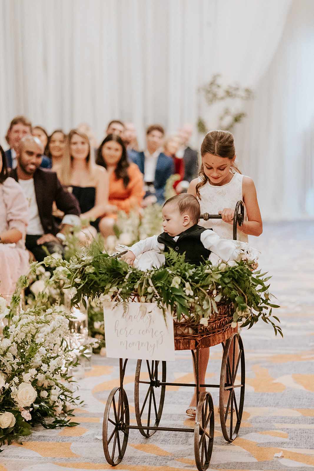 Flower Girl Pushing Ring Bearer in Wicker Wagon With Greenery and White Sign