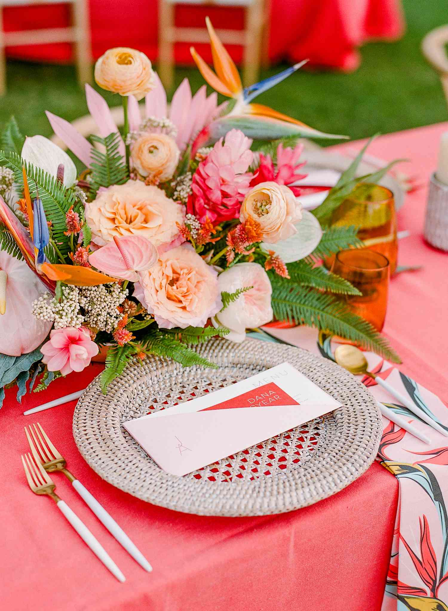 Pink tablescape with woven charger, tropical printed napkin, and pink, peach, and green floral arrangement