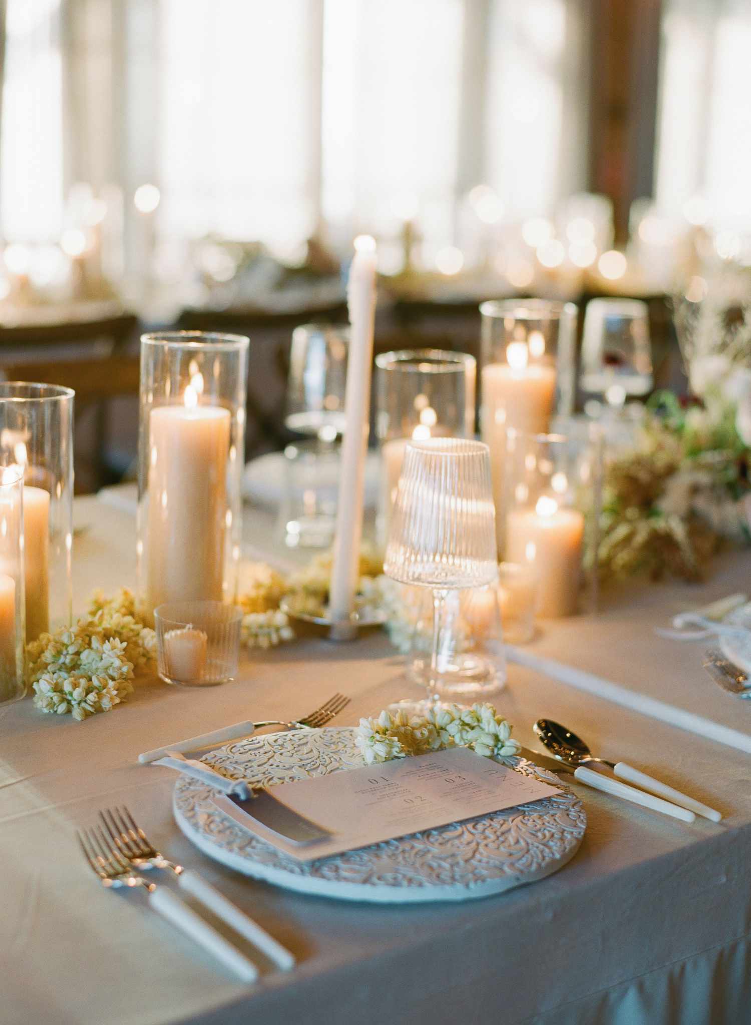 Place settings with white etched chargers, ribbed glassware, white florals, and candles