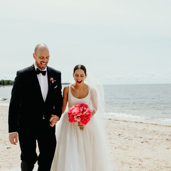A groom in a black suit and a bride in a white wedding dress carrying a pink bridal bouquet at a beach wedding venue.