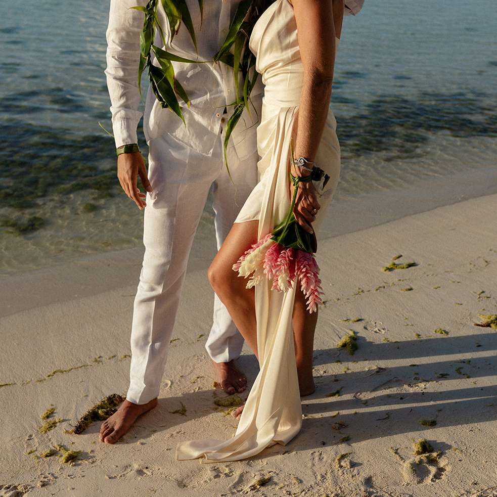 Man in white pants and shirt with woman in cream silk dress holding a bouquet of pink and white flowers standing on a tropical beach shore
