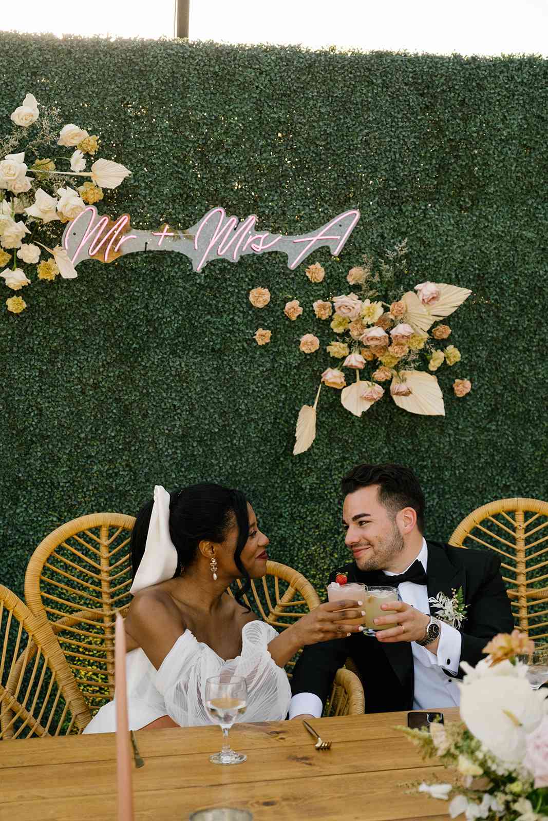 Bride and groom toast during dinner in front of pink neon "Mr + Mrs A" sign and neutral florals on hedge