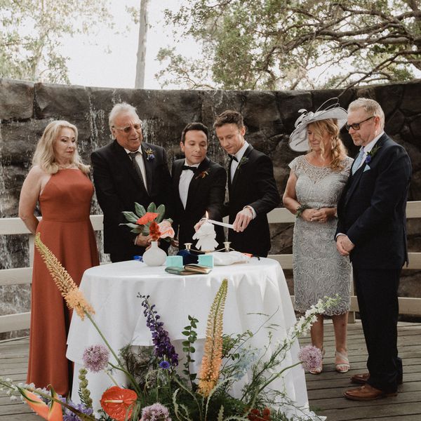 Two grooms lighting a large candle with taper candles, surrounded by family