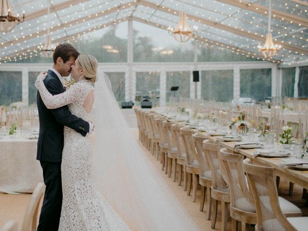 Bride and groom embracing, standing together under string lights in clear-top tent wedding venue with long wooden tables.