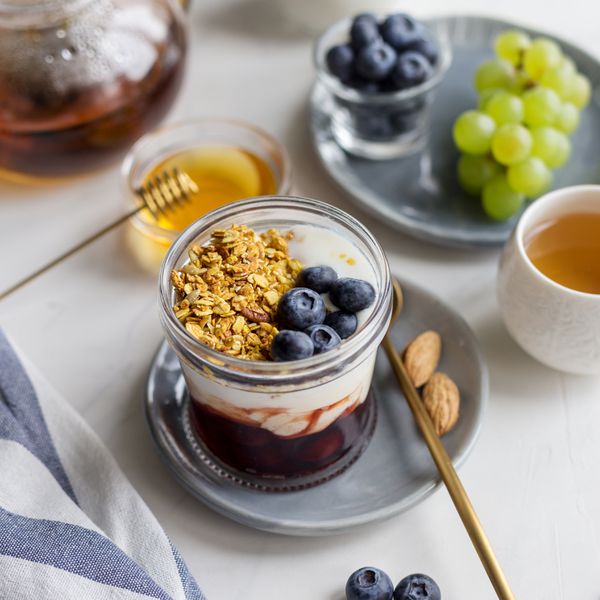 Summer breakfast in jar with granola, yogurt, jam and berries on gray dish with almonds, teapot and cup, napkin on white background.