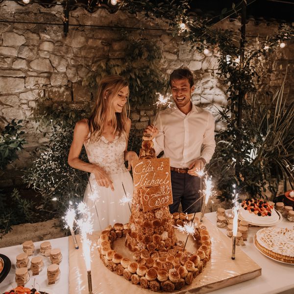 Bride in Lace Wedding Dress and Groom in Shirt and Pants Holding Sparklers Over Wedding Dessert Table With Variety of Small Treats