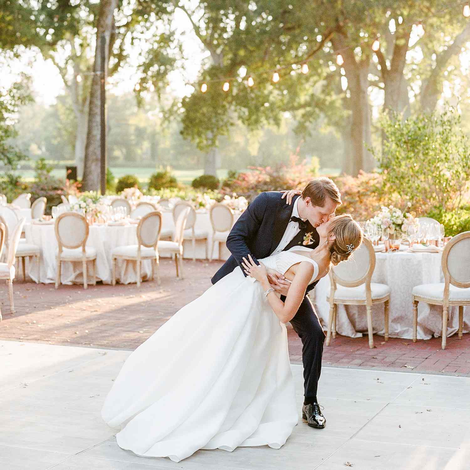 A bride and groom dancing on the dance floor in front of their outdoor wedding reception layout, consisting of white tables and chairs under trees and string lights.
