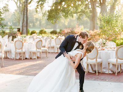 A bride and groom dancing on the dance floor in front of their outdoor wedding reception layout, consisting of white tables and chairs under trees and string lights.