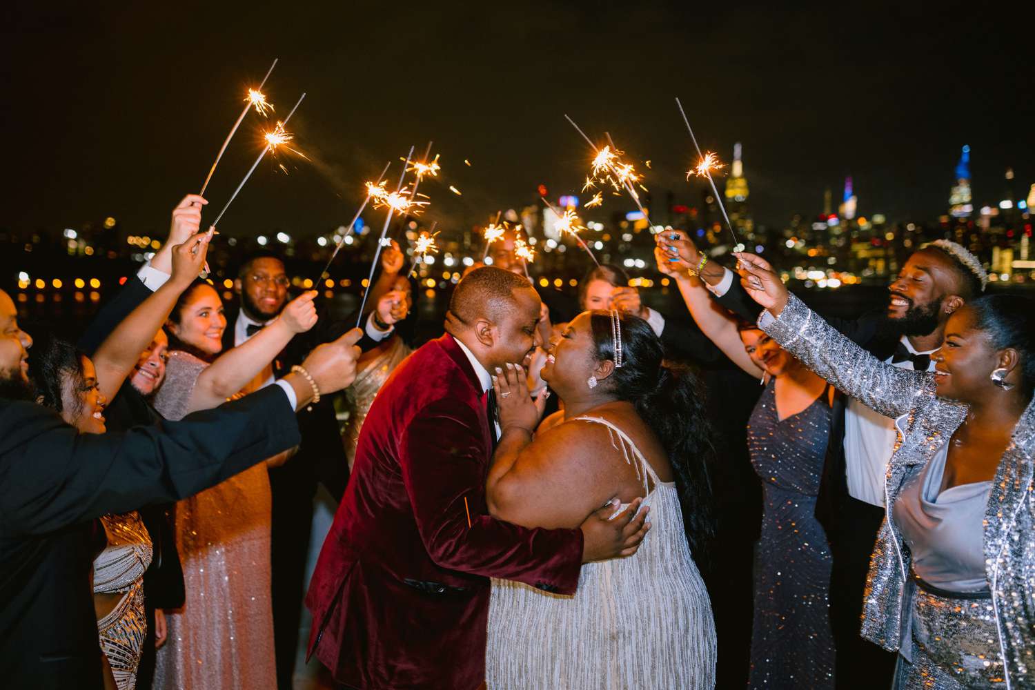 Bride and groom smiling at one another while their guests hold sparklers above them during nighttime in front of a city skyline backdrop