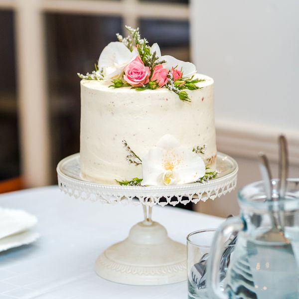 A single-tier white wedding cake with pink roses sitting on a delicate white cake stand.
