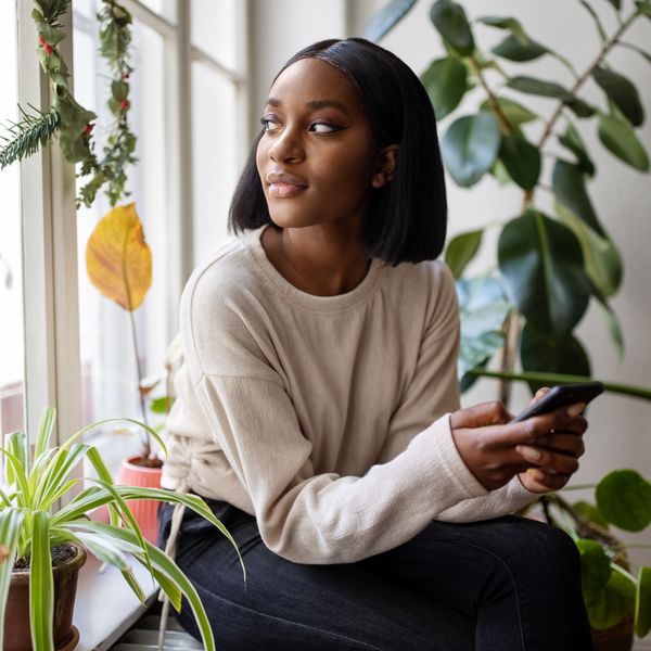 Woman Holding Phone in Apartment With Plants Stares Out the Window