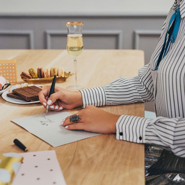 Woman in Striped Shirt Filling Out a Celebratory Card While Drinking a Glass of Champagne and Eating Chocolate