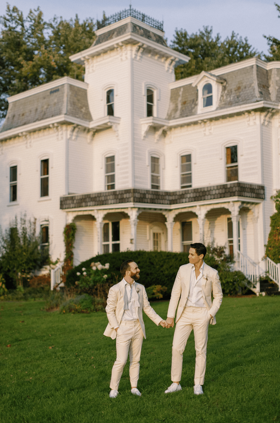 Two grooms in cream suits in front of white Victorian home wedding venue