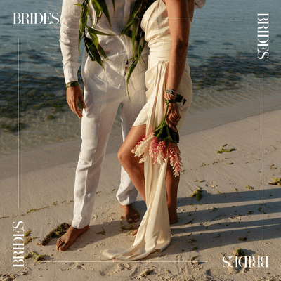 Man in white pants and shirt with woman in cream silk dress holding a bouquet of pink and white flowers standing on a tropical beach shore
